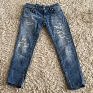 Dondup George 27\29 - Snygga blå jeans från dondup i modell Lambda, tillverkade i Italien. Jeansen har slitna detaljer och en klassisk femficksdesign med dragkedja och knapp. Raka ben och normal passform, perfekt för en avslappnad streetstil.