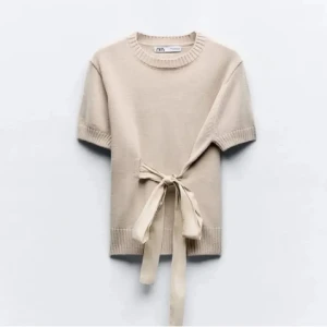 Beige stickad topp med knyt från Zara - Superfin beige stickad topp från Zara med korta ärmar och bred ribbad kant nertill. Snygg detalj med knytband i midjan som ger en unik look. Perfekt till jeans eller kjol för en chill men ändå stilsäker vibe.