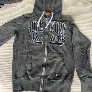Grå hoodie från Superdry Sport M - Snygg grå hoodie från Superdry Sport med dragkedja, huva med snörning och stora tryckta bokstäver framtill. Mjuk insida och känguruficka gör den extra bekväm. Perfekt för chill eller träning.