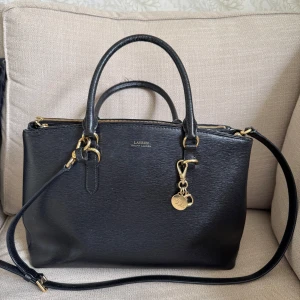 Svart handväska från Lauren Ralph Lauren - Snygg svart handväska från Lauren Ralph Lauren i skinn med guldfärgade metalldetaljer och dubbla dragkedjor. Väskan har både handtag och axelrem, samt en dekorativ RL-berlock. Kommer med original dustbag och etiketter. Modell double zip large satchel. Perfekt storlek, rymmer datorn. Bra begagnat skick. 