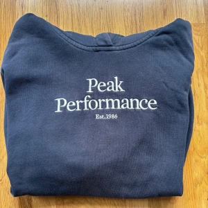 Mörkblå Peak Performance hoodie - Mörkblå hoodie från Peak Performance med broderad vit logga och text 'Est. 1986' på bröstet. Klassisk känguruficka framtill, huva och ribbade muddar. Perfekt för chill dagar eller när du vill ha en clean och stilren look.