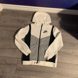 Nike hoodiejacka i vitt och grått - Nike hoodiejacka i storlek L med dragkedja framtill. Jackan har en snygg färgblock-design i vitt, grått och svart, samt en broderad Nike-logga på bröstet. Tillverkad i mjuk bomullsmix och har huva och långa ärmar. Perfekt för en sportig och chill stil.