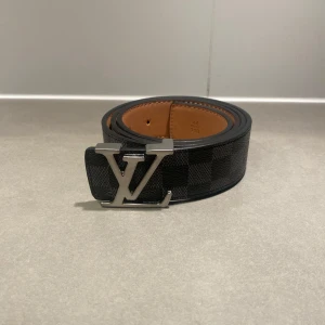 Svart LV bälte - Snyggt Louis Vuitton bälte i svart och grått med klassiskt rutigt Damier Graphite-mönster. Bältet har en stor silverfärgad LV-logga som spänne och är gjort i exklusivt skinn med brun insida. Perfekt accessoar för att lyfta din outfit.
