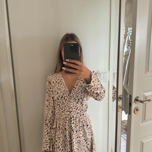 Beige blommig klänning med v-ringning - Supersöt beige klänning med svart blommönster och v-ringning. Klänningen har lång ärm och är lite tajtare i midjan för en snygg passform. Perfekt för vår och sommar! Aldrig använd