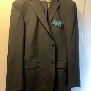 Kostym - Väst, Kavaj, Byxor - Snygg svart kostym från SIR med diskreta ränder. Inklusive kavaj med blå näsduk, väst och byxor. Klassisk passform och stilren look. Materialmix av ull och polyester. Kavajen är från märket Sir, byxor och kavaj är från Batistini. Allt använt enbart en gång.