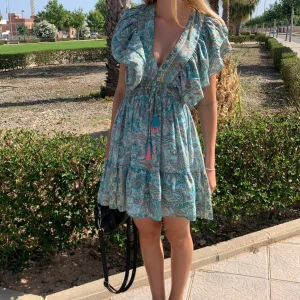 Blå paisley kortklänning med volang - Supersöt kortklänning i blått och turkost paisleymönster med volangdetaljer och djup v-ringning. Klänningen har knytband med tofsar i midjan. Köpt i Spanien och endast använd 1 gång🥰🥰storleken passar XS-M skulle jag säga.