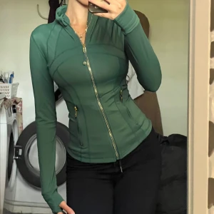 Grön Define Jacket från Lululemon - Supersnygg grön Define Jacket från Lululemon med guldiga dragkedjor och figurnära passform. Jackan har långa ärmar med tumhål, hög krage och två fickor framtill. Perfekt för träning eller chill dagar.