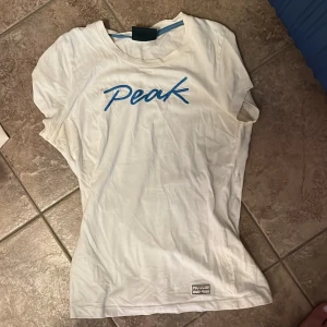 Vit Peak Performance t-shirt med blått tryck - Vit t-shirt från Peak Performance med broderad blå text 'Peak' på bröstet. Klassisk rund halsringning med blå detalj på insidan. Liten etikett nertill med logga. Skön passform och perfekt basic-plagg för din garderob.