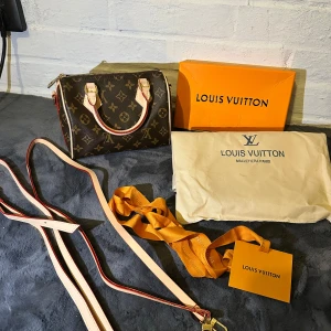 Louis Vuitton Monogram handväska - Säljer en klassisk Louis Vuitton handväska i brunt monogrammönster med beige och röda detaljer. Väskan har guldfärgade metalldetaljer, dragkedja och avtagbar axelrem. Ikonisk design med LV-logga och blommor, perfekt för dig som gillar lyxiga accessoarer.