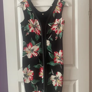 Blommig klänning från Guess - Säljer en svart ärmlös klänning från Guess med blommor i rött, vitt och grönt. Klänningen har en tight passform och är perfekt för sommarens tillfällen. Snygg och stilren design med rund halsringning.