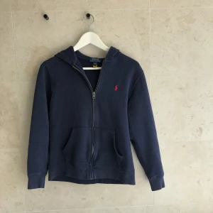Marinblå hoodie med dragkedja från Polo Ralph Lauren - Mörkblå hoodie från Polo Ralph Lauren med dragkedja och huva. Klassisk röd logga broderad på bröstet och två fickor framtill. Perfekt för en avslappnad stil.Nypris 1200kr vid snabb affär 500kr