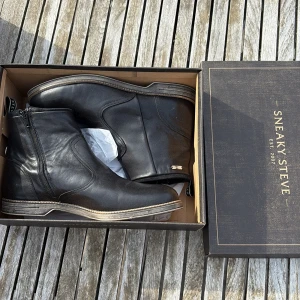 Svarta boots från Sneaky Steve - Säljer ett par svarta boots från Sneaky Steve i skinn med dragkedja på sidan. Snygg kontrastsöm runt sulan och klassisk design med rund tå. Perfekta för dig som gillar stilrena skor med lite edge.