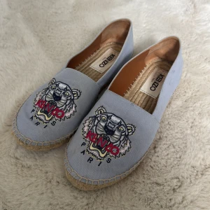 Ljusblå espadrillos från Kenzo med tigermotiv - Säljer ett par grå espadrillos från Kenzo med det ikoniska broderade tigermotivet och texten 'KENZO PARIS' framtill. Skorna har rund tå och flätad sula i klassisk espadrillostil. Perfekta för en trendig och avslappnad look.