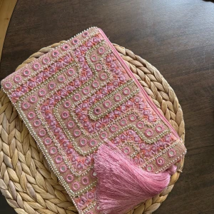 Broderad rosa kuvertväska med tofs - Superfin kuvertväska i rosa med broderade detaljer i guld och pärlor. Väskan har ett snyggt geometriskt mönster på framsidan och en stor rosa tofs vid dragkedjan. Perfekt storlek för mobilen och det viktigaste. Mjuk bomullskänsla och helfärgad baksida.