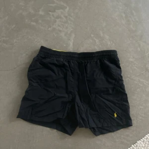 Svarta badshorts från Polo Ralph Lauren - Svarta badshorts från Polo Ralph Lauren med gul broderad logga på benet. Resår och snörning i midjan, insida i vitt meshfoder och gul insida på linningen. Perfekta för stranden eller poolen.