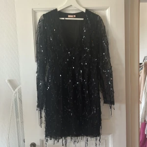 Svart paljettklänning med v-ringning - Säljer en svart klänning med v-ringning och långa ärmar, täckt av glittriga paljetter. Perfekt för dig som vill sticka ut på festen! Klänningen har en kort längd och ger en riktigt glam känsla.