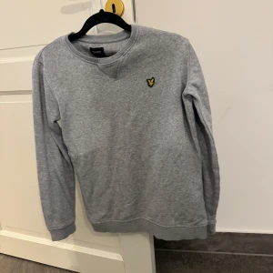 Grå sweatshirt från Lyle & Scott - En stilren grå sweatshirt från Lyle & Scott med klassisk rund hals och det ikoniska gula örnloggan broderad på bröstet. Tröjan har långa ärmar och ribbade muddar vid ärmslut och nederkant. Perfekt för en chill och avslappnad look.