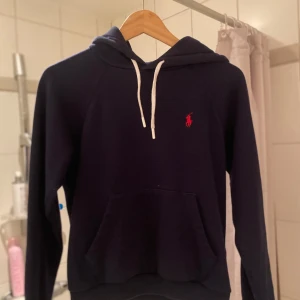 Mörkblå hoodie från Polo Ralph Lauren - Säljer en snygg mörkblå hoodie från Polo Ralph Lauren med det klassiska röda logotypen på bröstet. Tröjan har en känguruficka och vita dragsnören vid huvan. Perfekt för en avslappnad stil. Storlek S passar runt 160-165. Skick 9/10 använd fåtal gånger inga defekter. Hör av er vid frågor!