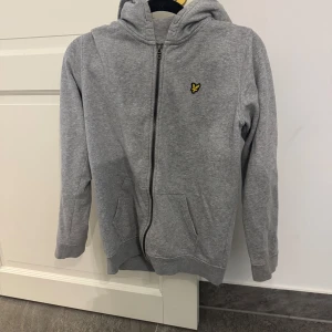 Grå hoodie från Lyle & Scott - Snygg grå hoodie från Lyle & Scott med dragkedja och klassisk gul logga på bröstet. Tröjan har huva, två fickor framtill och är tillverkad i mjuk bomullsmix. Perfekt för en chill och avslappnad stil.
