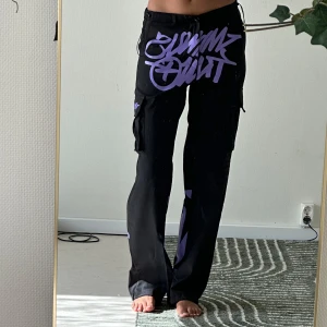Svarta cargobyxor med lila tryck - Säljer ett par svarta cargobyxor med stora lila tryck och grafiska detaljer. Byxorna har vida ben och flera fickor på sidorna. Perfekta för dig som gillar streetwear och vill sticka ut med coola detaljer.