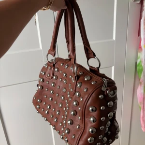 Brun handväska med nitar - Cool brun handväska med stora silvriga nitar över hela väskan. Väskan har två handtag och är tillverkad i ett material som liknar skinn. Perfekt för dig som vill sticka ut med en edgy accessoar.
