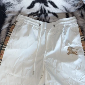 Vita shorts med rutiga sidopaneler och broderad logga - Snygga vita shorts/badbyxor med klassiska Burberry-rutiga sidopaneler. Kan ta flera bilder bara fråga ifall intresse. SOMMAR TIDEN ÄR FRAME