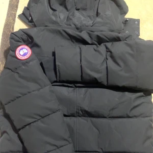 Svart dunjacka från Canada Goose M - Säljer en svart dunjacka från Canada Goose med huva och avtagbar pälskant. Jackan har klassisk patch på ärmen, dragkedja och knappar framtill samt rejäla muddar vid ärmslut. Perfekt för kalla vinterdagar och riktigt snygg streetstil.