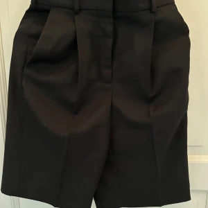 Svarta kostymshorts Acne Studios - Stilrena svarta kostymshorts från Acne Studios med hög midja och pressveck. Klassisk design med bälteshällor, knappar och fickor bak. Materialmix av ull och polyester ger en dressad känsla. Perfekta för en clean och modern look.