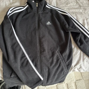 Fleecejacka Adidas - Svart fleecejacka från Adidas med dragkedja och klassiska vita ränder längs ärmarna. Jackan har hög krage, två fickor och Adidas-logga på bröstet. 