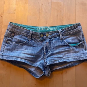 Blå jeansshorts från Cross-X - Säljer ett par blå jeansshorts från Cross-X med dubbla knappar framtill och dekorativa nitar på bakfickorna. Shortsen har klassisk femficksdesign och turkos insida i midjan. Perfekta för varma dagar!