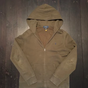 Olivgrön hoodie från Polo Ralph Lauren - Säljer en olivgrön hoodie från Polo Ralph Lauren med dragkedja framtill och två fickor. Tröjan har en liten broderad logga i blått på bröstet och är tillverkad i mjukt material. Perfekt för en avslappnad stil. Skicka kring fler frågor. Lite osäker kring storlek. Jag är dock ca 175 cm lång och den är lite kort för mig enligt mig 