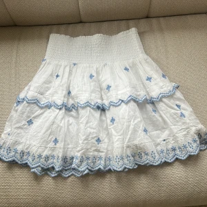 Vit kjol med blå broderi från Neo Noir - Supersöt vit kortkjol från Neo Noir med blå broderade detaljer och volangkant. Kjolen har smock i midjan och är gjord i ett lätt bomullstyg, perfekt för varma dagar. Broderierna ger en romantisk vibe och volangerna gör den extra gullig.Köpt från Jackie.