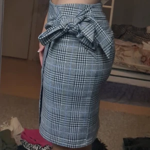 Rutig omlottkjol med rosett - Snygg rutig kjol i omlottmodell med stor rosett i midjan. Kjolen har en asymmetrisk nederkant och klassiskt rutmönster i svart, vitt och grått. Perfekt för dig som gillar en trendig och stilren look.