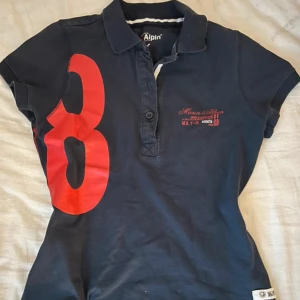 Marin Alpin mörkblå pikétröja med röd detalj - Mörkblå pikétröja från Marin Alpin med stor röd siffra 8 på både fram- och baksida. Röd broderad logga på bröstet och ryggen samt röda detaljer vid nacken. Klassisk krage och korta ärmar. Perfekt för en sportig look.