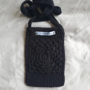 Svart virkad axelväska från ElindaDesign - Säljer en svart virkad axelväska från ElindaDesign. Väskan har ett unikt mönster framtill och en bred axelrem. Perfekt för att bära mobilen eller småsaker. Enkel och stilren design med handgjord känsla.