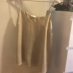 Beige linnetopp från C - Säljer en stilren beige linnetopp från Cubus i storlek S. Toppen har tunna axelband och v-ringad halsringning, tillverkad i ett glansigt, mjukt material som ger en lyxig känsla. Perfekt att styla med jeans eller kjol.
