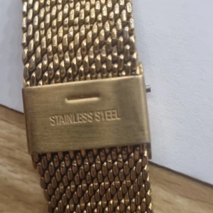 Guldfärgad klocka från Kompass - Stilren klocka från Kompass med guldfärgat mesharmband i rostfritt stål och vit urtavla. Klockan har minimalistisk design med tunna visare och index. Baksidan är graverad med kompassros och information om vattentäthet (3 ATM).