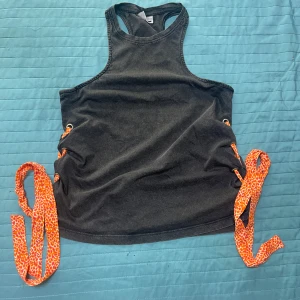 Mörkgrå linnetopp med orange snörning - Cool mörkgrå linnetopp med breda axelband och snygga orange snören i leopardmönster som går genom metallöljetter på sidorna. Toppen har ärmlös design och  perfekt för en edgy och trendig look.