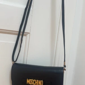 Svart axelväska från Moschino - Snygg svart axelväska från Moschino med guldiga bokstäver framtill. Väskan har lång, smal axelrem och stilren design. Perfekt för dig som gillar klassiska accessoarer med en touch av lyx. Tillverkad i slätt skinn.