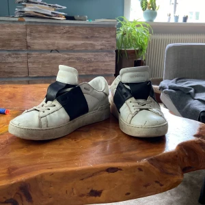 Valentino Open sneakers vit/svart skinn - Snygga Valentino Open sneakers i vitt skinn med bred svart läderdetalj över mitten. Klassisk rund tå, snörning och coola nitar baktill på sulan. Perfekta för dig som gillar exklusiva och trendiga sneakers med unik design.