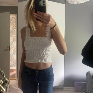 Sommar topp  - Säljer en söt vit croppad linnetopp med breda axelband och volangkant nertill. Toppen har smockad resår över hela och är perfekt för varma dagar.