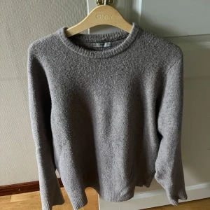 Grå stickad tröja från Mango  - Mysig grå stickad tröja från Mango med rund halsringning och långa ärmar. Perfekt för kyliga dagar och enkel att matcha med jeans eller kjol. Tröjan har en klassisk, avslappnad passform och ribbade muddar vid ärmslut och nederkant.