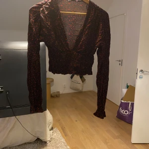 Svart blommig omlottopp med smock - Supersöt svart omlottopp med små röda blommor och smockad midja samt ärmslut. Toppen har v-ringning, långa ärmar och croppad passform. Perfekt för dig som gillar romantiska detaljer och vill ha något som sticker ut lite extra.