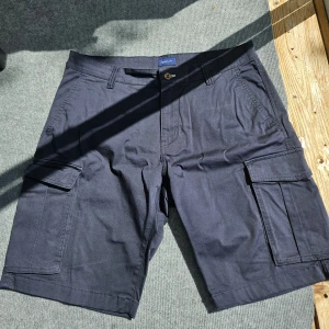Mörkblå cargo shorts från GANT - Snygga mörkblå cargo shorts från GANT med flera fickor på sidorna och bak. Klassisk design med avslappnad passform och knappgylf. Perfekta för sommardagar när du vill ha både stil och funktion.