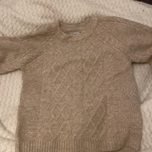 Beige stickad tröja från 157 - Mysig beige stickad tröja från 157 med klassiskt flätmönster framtill och längs ärmarna. Tröjan har rund halsringning och ribbade muddar vid ärmslut och nederkant. Perfekt för kyliga dagar!