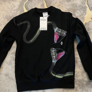 Svart tröja med ormmotiv Marcel - Svart långärmad tröja från Marcel County of Milan med ett fett tryck av två ormar med öppna gap i grönt, grått och rosa. Crewneck-modell i mjuk bomull, perfekt för dig som vill sticka ut med en edgy vibe. Strl S