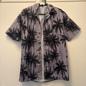 palmmönstrad skjorta - Snygg kortärmad skjorta med svart palmmönster. Skjortan har klassisk krage, knäppning framtill och är tillverkad i ett lätt syntetmaterial. Perfekt för dig som vill sticka ut med tropiska vibes.