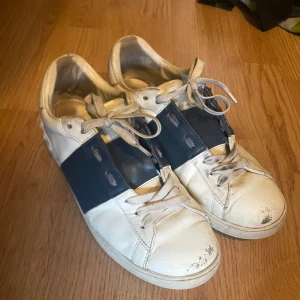 Valentino Open sneakers vit/blå - Snygga Valentino Open sneakers i vitt skinn med bred mörkblå rem över mitten. Klassisk rund tå, vita snören och platt sula. Diskret Valentino-logga på sulan. Perfekta för dig som gillar stilrena och exklusiva sneakers. Hjälper en polare att sälja då han bara vill ha lite snabba pengar!