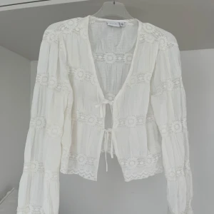 Vit blus med spetsdetaljer och knyt - Säljer en romantisk vit blus med långa ärmar, spetsinslag och två knyt framtill. Blusen har vackra broderade detaljer och en luftig känsla, perfekt för vår och sommar.