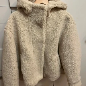 Beige teddyjacka med huva från ZARA - Mysig beige teddyjacka från ZARA i nyskick med huva och dragkedja. Jackan har lång ärm och två fickor framtill. Perfekt för kyliga dagar och ger en trendig look med sin fluffiga struktur.🥰🥰 